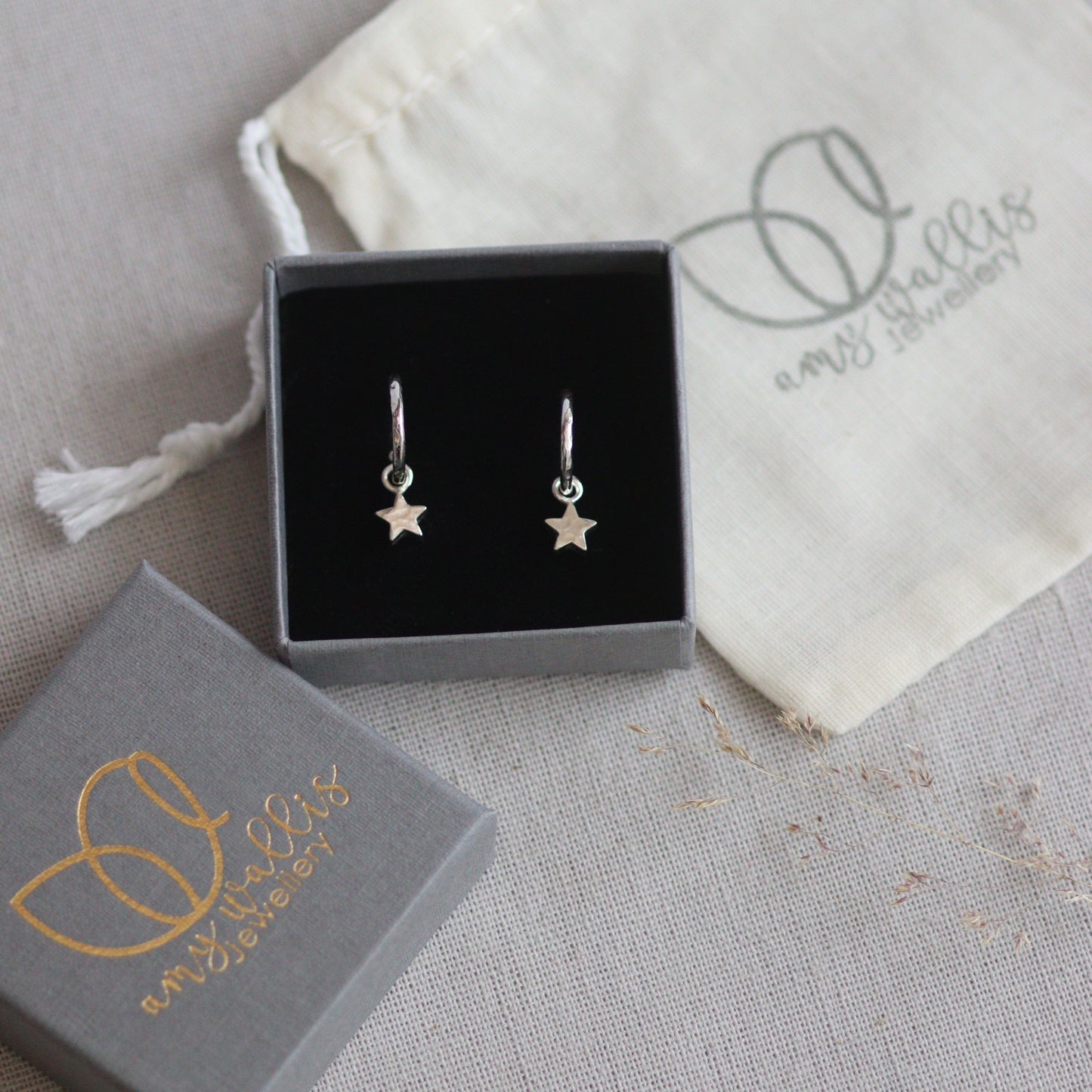 Silver star huggie hoop earrings in a box with a branded pouch on a gray surface