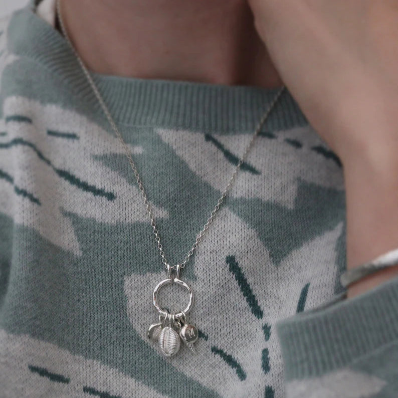A modern silver charm necklace being worn with a combination of seashell charms and initial pendants on the keeper ring against a soft green jumper.