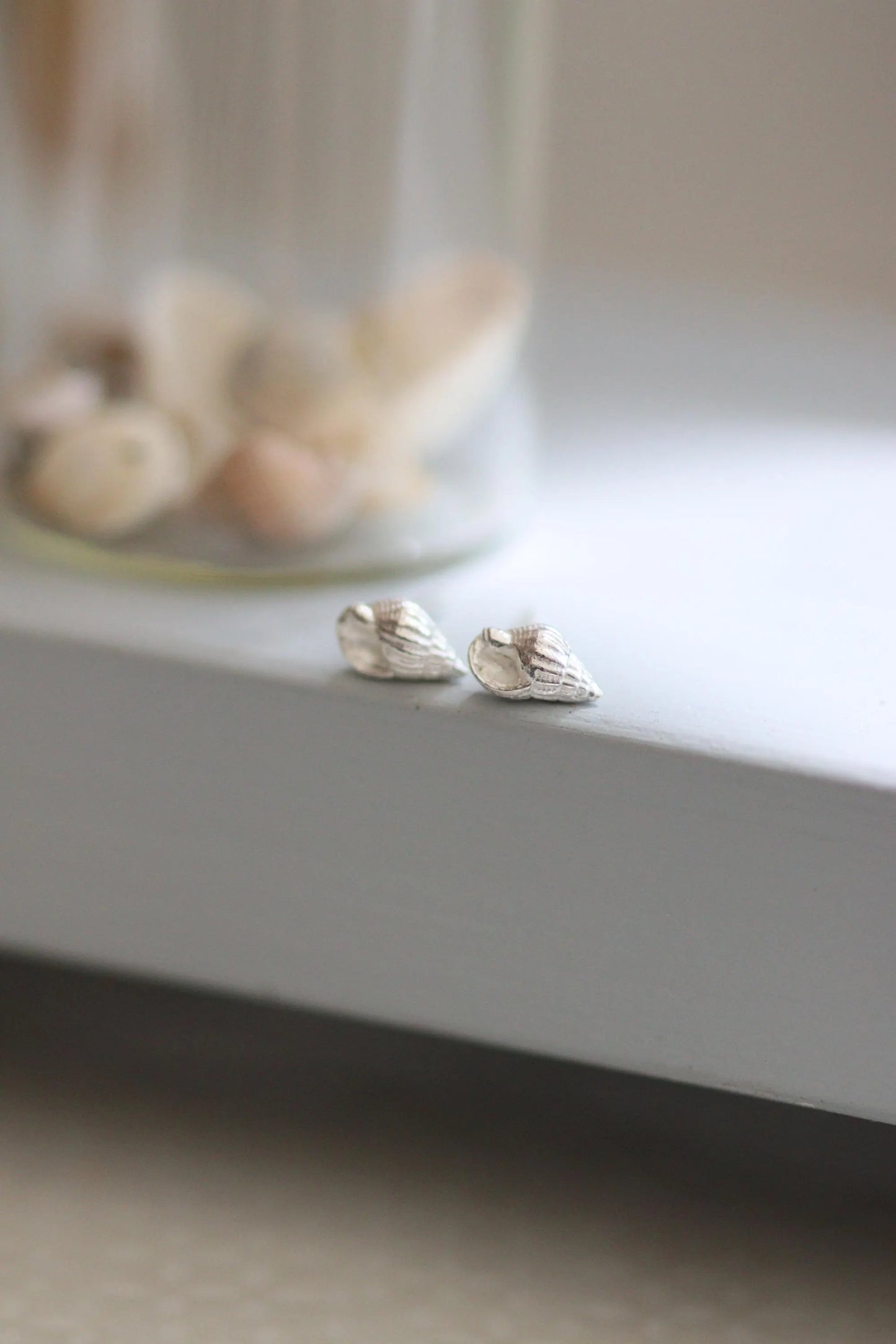Small solid silver seashell earrings on a grey shelf in front of a bottle of shells.