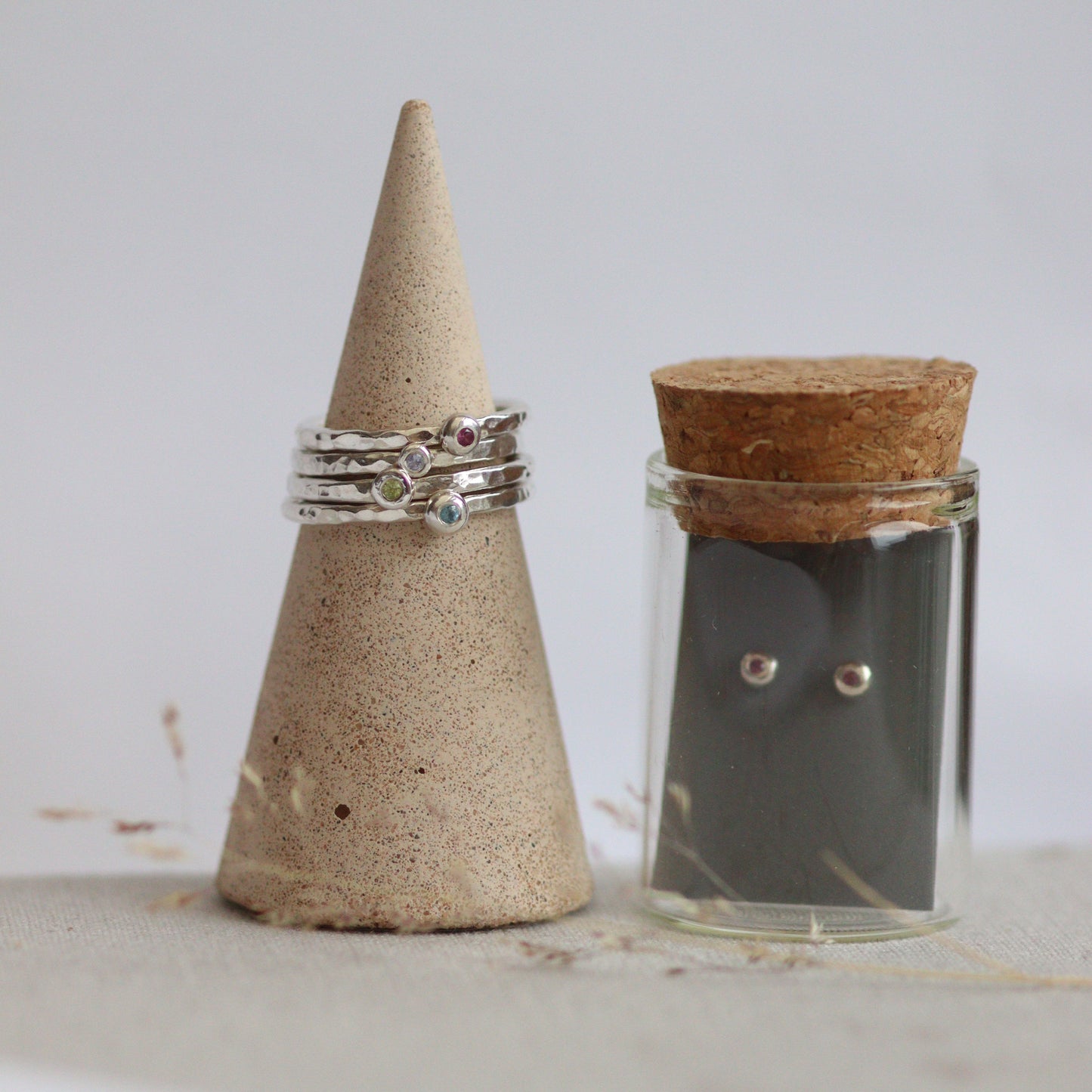 Stack of hammered silver stacking rings with coloured birthstones next to a glass bottle containing tiny silver birthstone studs.