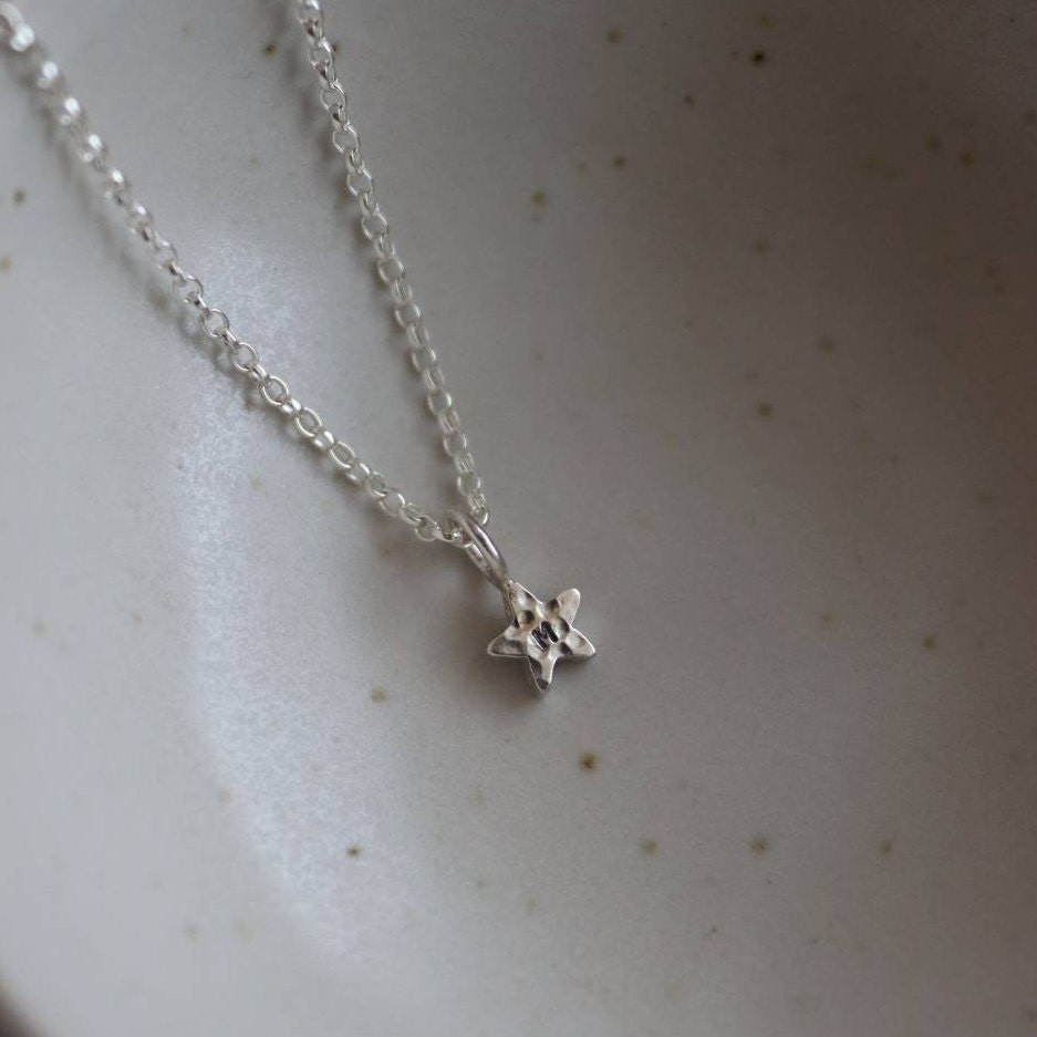 Hammered silver star pendant with an initial stamped on a fine silver chain on a light beige background.