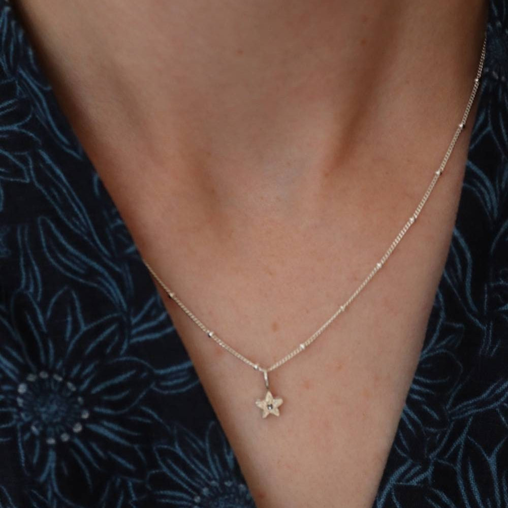 A silver star charm with initial personalisation on a fine beaded silver chain worn on a model in a blue v-neck.