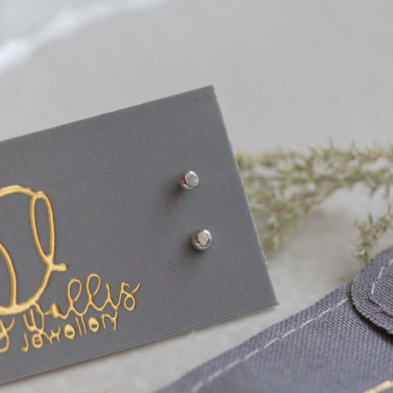 Pair of silver stud earrings on a grey card with gold text, placed on a light surface.