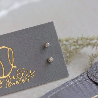 Pair of silver stud earrings on a grey card with gold text, placed on a light surface.