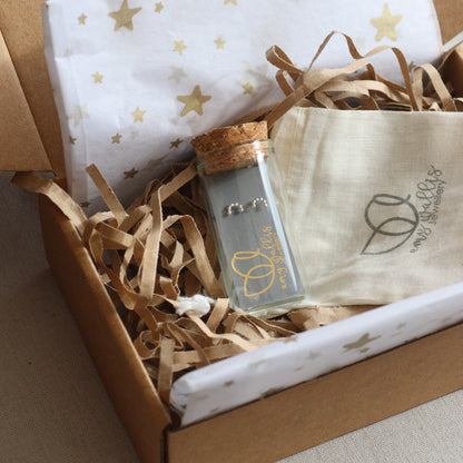 Product packaging with a bottle and silver rainbow earrings on star-patterned tissue inside a cardboard box.