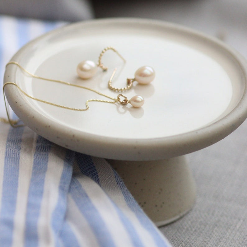Gold pearl hoop earrings and a classic minimalist small pearl drop necklace on a small ceramic stand with a striped fabric background