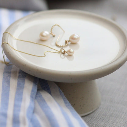 Gold pearl hoop earrings and a classic minimalist small pearl drop necklace on a small ceramic stand with a striped fabric background