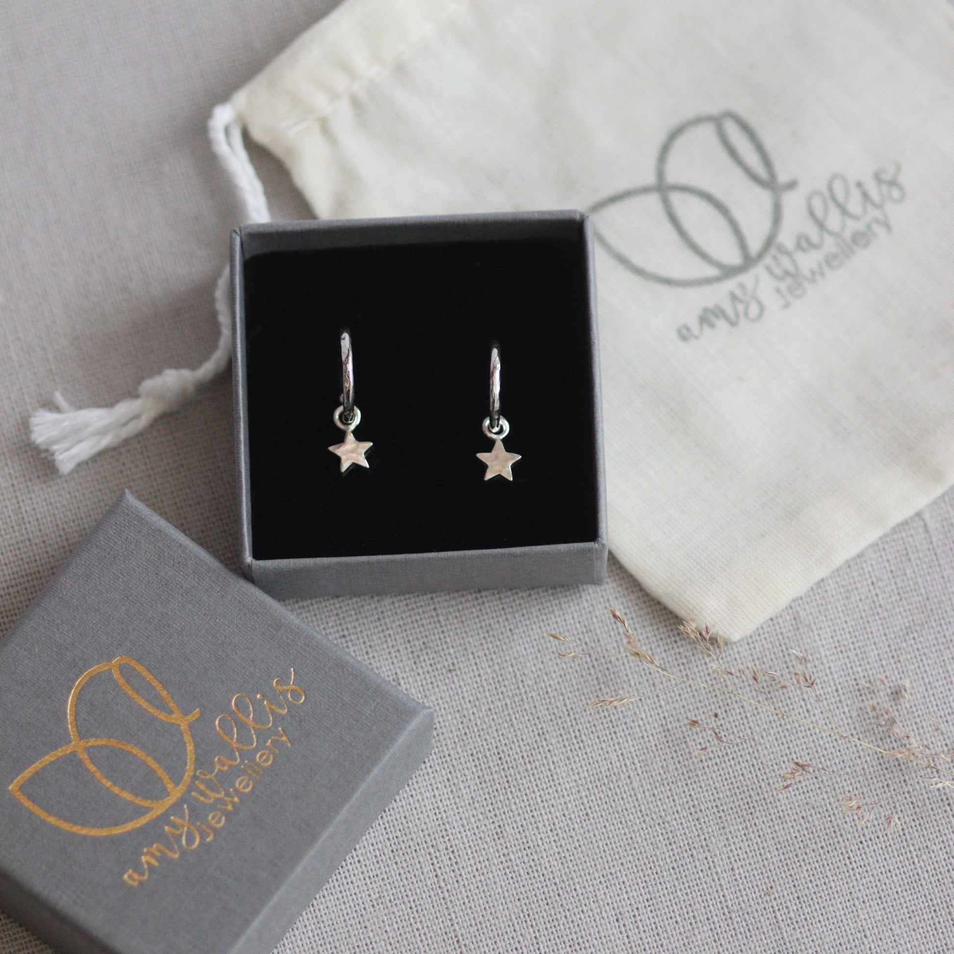 Silver star huggie hoop earrings in a box with a branded pouch on a gray surface