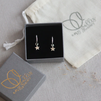 Silver star huggie hoop earrings in a box with a branded pouch on a gray surface