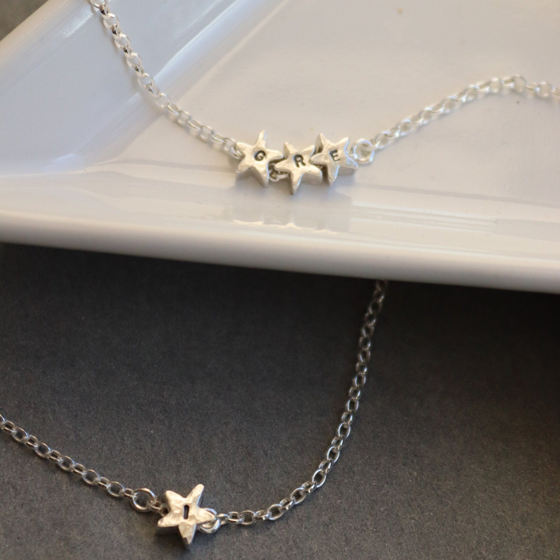 Silver star necklace with initials on the stars on a white dish with a grey surface