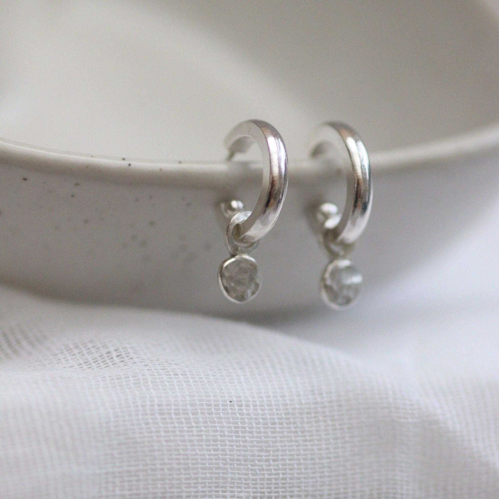 A pair of silver huggie hoop earrings with shimmering confetti dangles, placed on a white surface with a textured background.