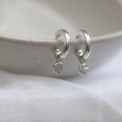 A pair of silver huggie hoop earrings with shimmering confetti dangles, placed on a white surface with a textured background.