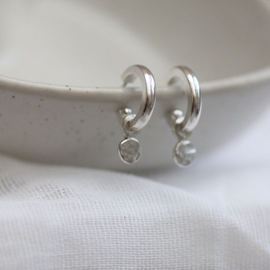 A pair of silver huggie hoop earrings with shimmering confetti dangles, placed on a white surface with a textured background.