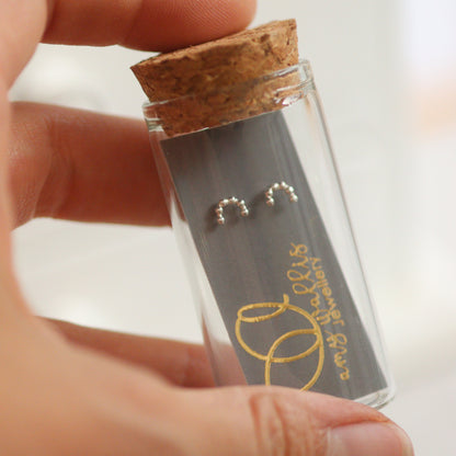 Small glass tube with cork lid containing silver rainbow earrings, held by a hand against a neutral background