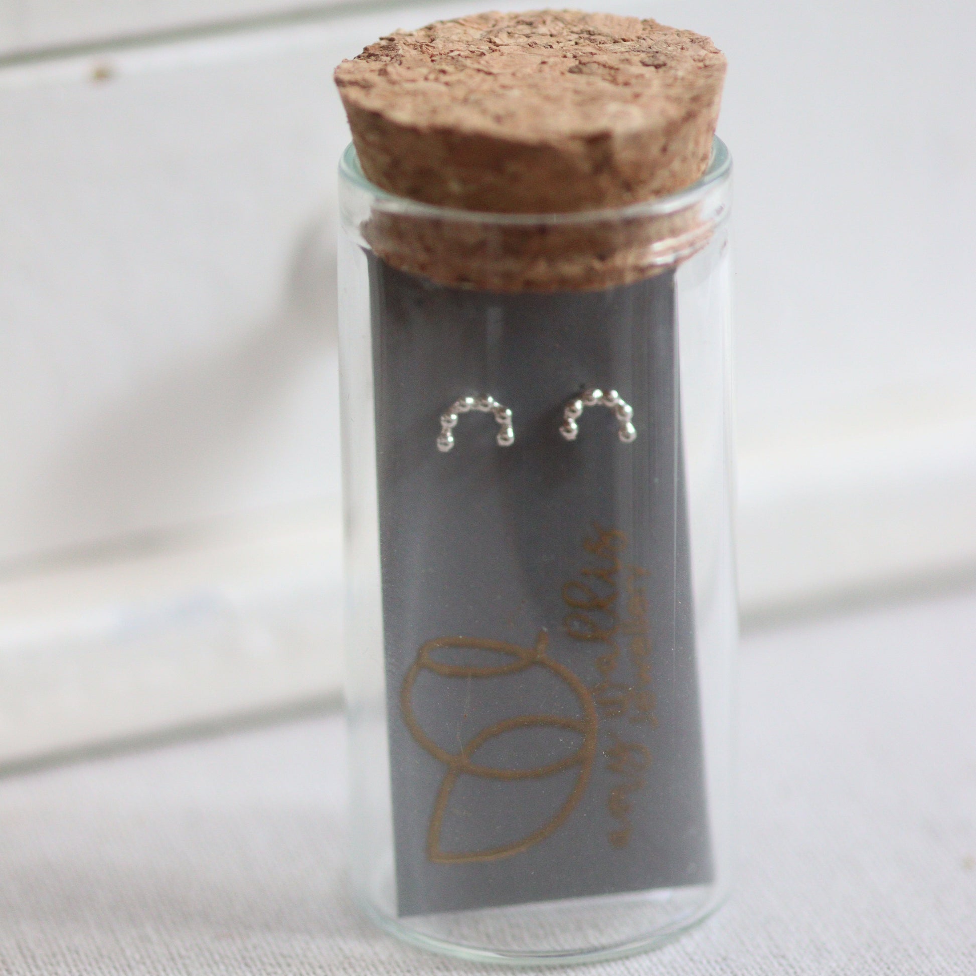 Glass vial with cork lid containing silver minimalist rainbow earrings on a white background