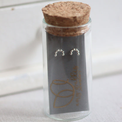 Glass vial with cork lid containing silver minimalist rainbow earrings on a white background
