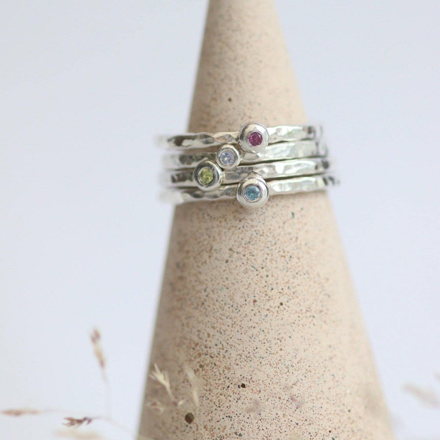 Stack of silver rings with colourful gemstones on a textured cone-shaped stand against a light background