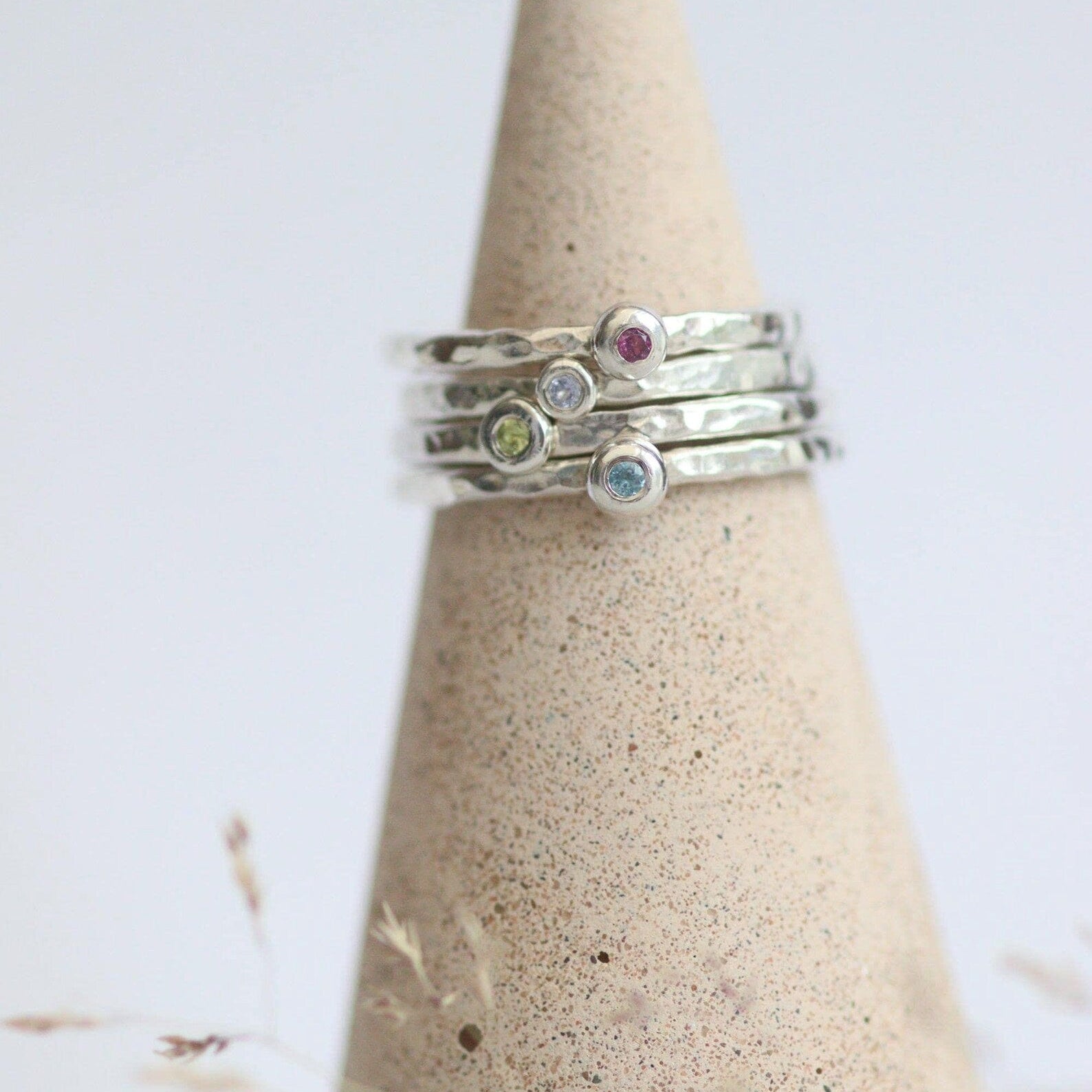 Stack of silver rings with colourful gemstones on a textured cone-shaped stand against a light background