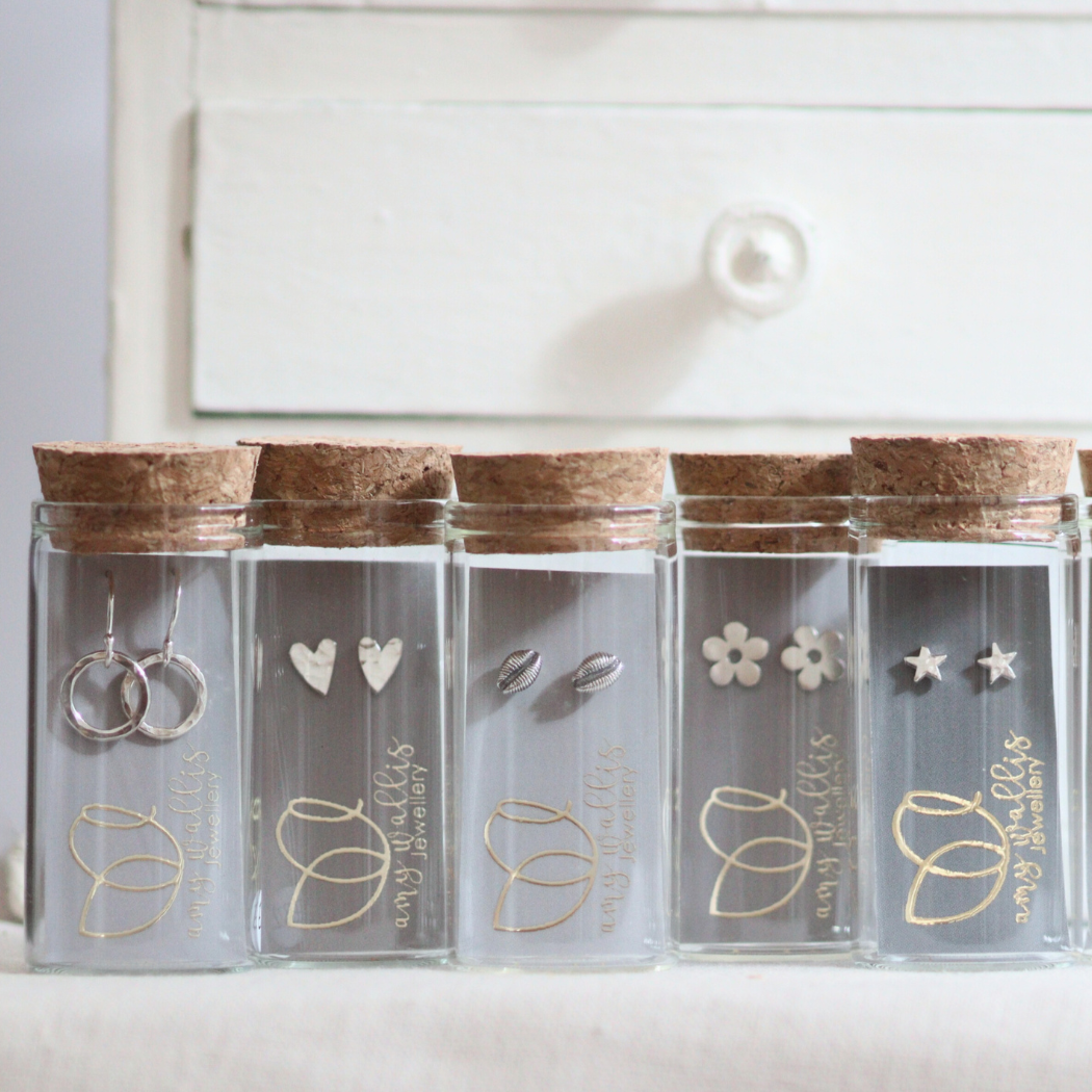 A row of various handmade silver stud earrings packaged in unusual glass bottles with corks and grey branded cards.