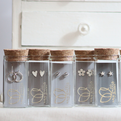 A row of various handmade silver stud earrings packaged in unusual glass bottles with corks and grey branded cards.