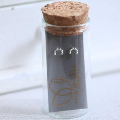 Glass jar with cork lid containing silver arc earrings on a white wooden surface