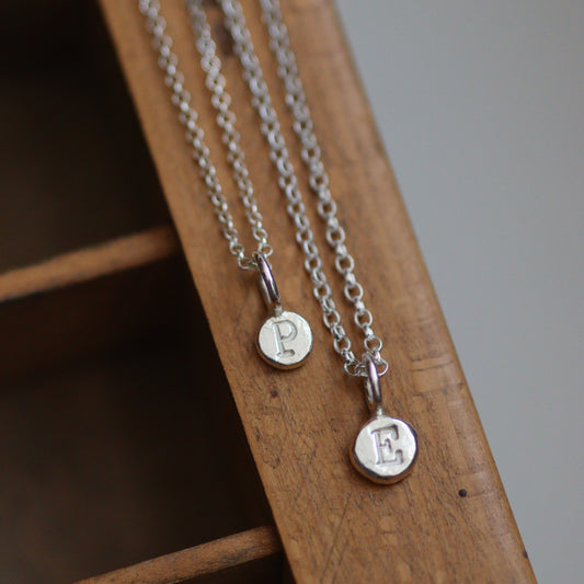 Two silver personalised initial necklaces with round pendants on a wooden surface
