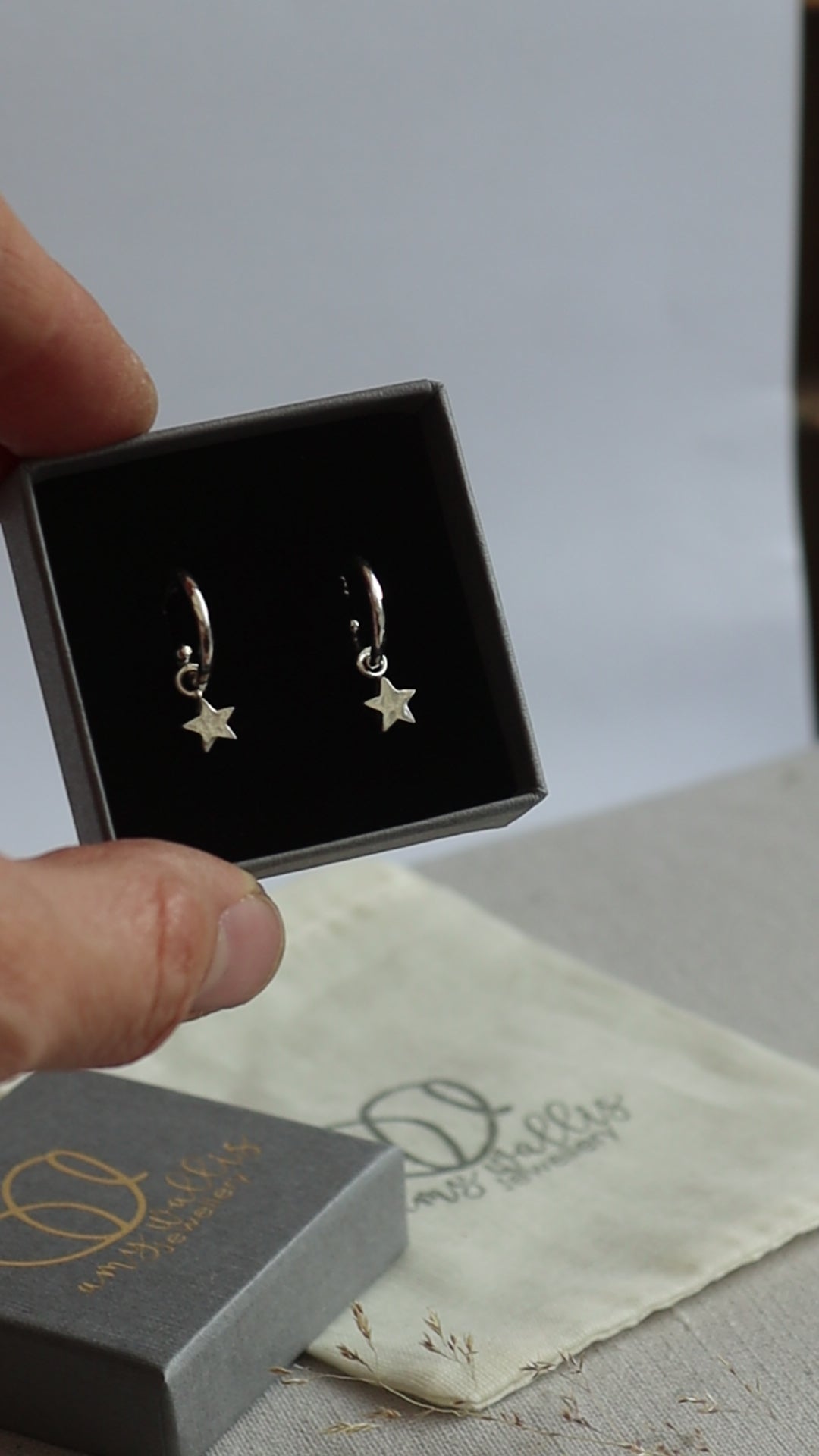 Silver hammered huggie hoops with star dangles in a grey branded jewellery box.