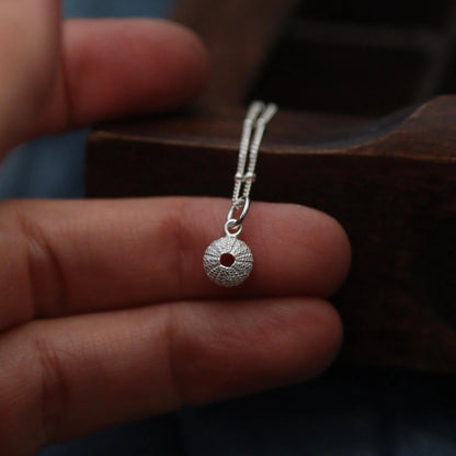 silver patterned sea urchin pendant on a beaded chain in front of a letterpress drawer