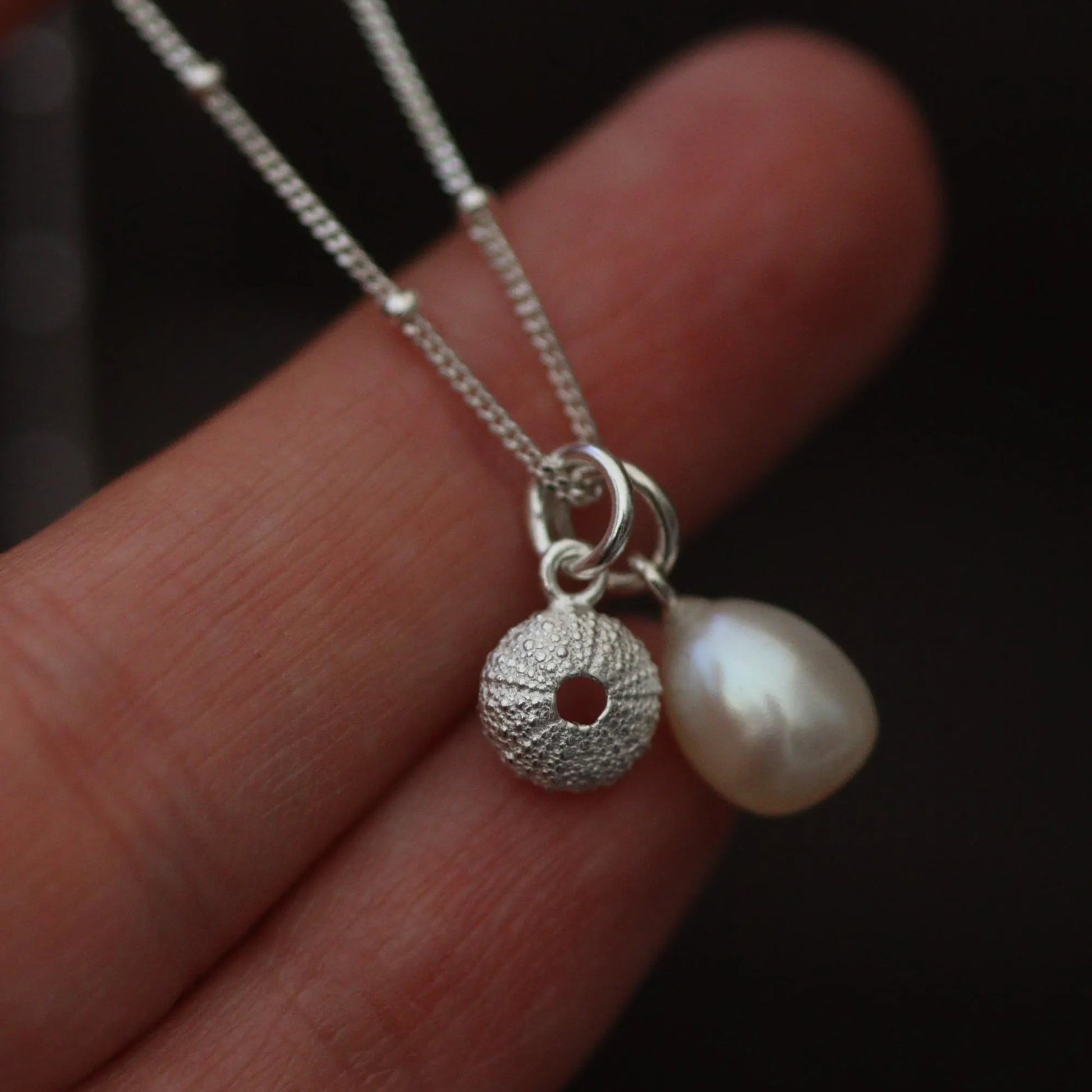 silver sea urchin charm on a chain with a pearl pendant being held against a hand