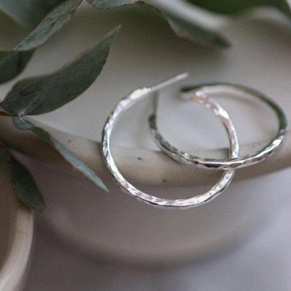 Large hammered hoop earrings, silver colour on a light background with eucaluptus.