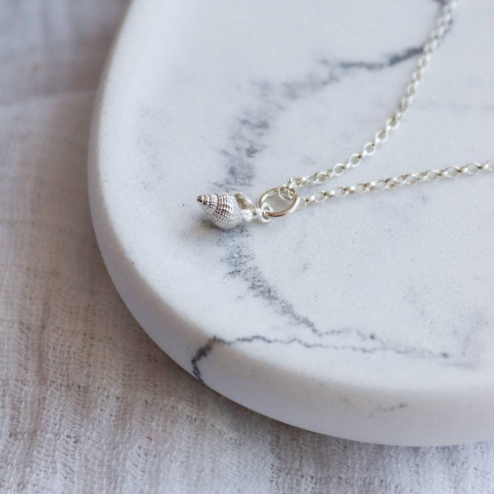 A tiny mudeford whelk cast in solid silver and strung on a silver chain. The necklace is on a marble dish.