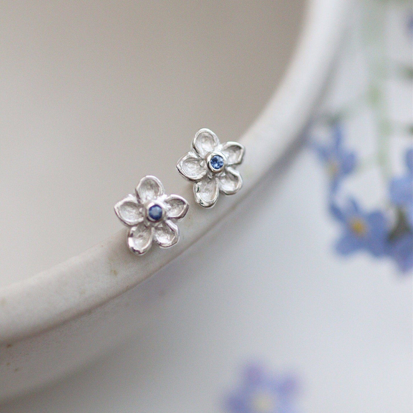 silver forget me not stud earrings with sapphire centre stones alanced on the edge of a bowl