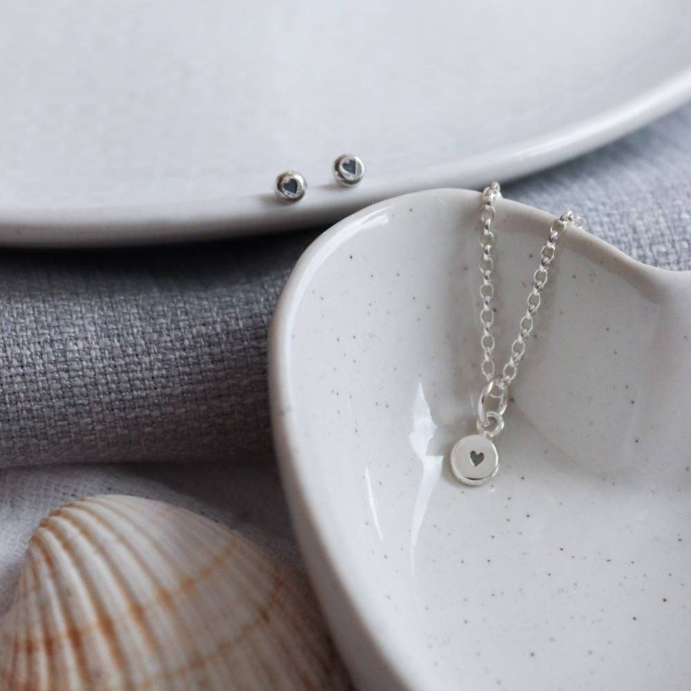 A pair of heart stamp studs and a silver heart stamp necklace draped in a bowl.