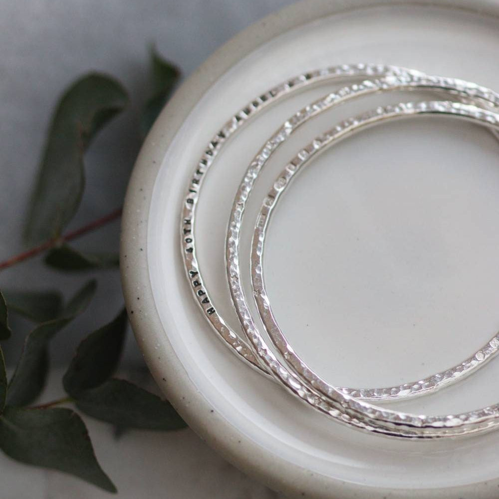 3 solid silver bangles lie on a dish, they hav a bright hammered finish and words of personalisation stamped around the bangle.