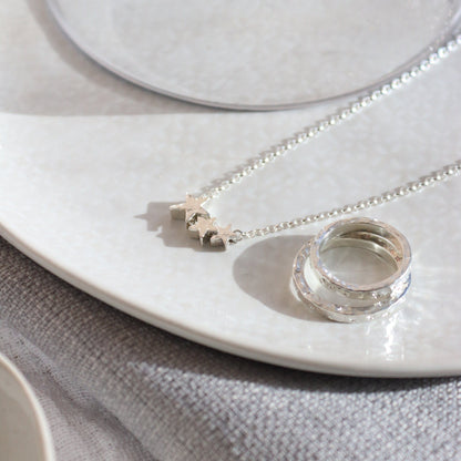 a silver necklace with 3 silver star beads graduating in size from big to small next to a stack of hammered rings with the same finish.